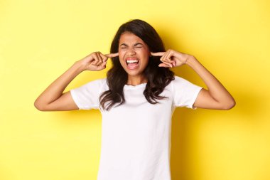 Image of pissed-off african-american girl, cant stand loud bothering noise, shut ears and screaming annoyed, standing over yellow background