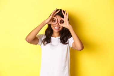 Mutlu ve komik Afro-Amerikan kızı tasviri, parmak gözlüğü yapıyor ve onlara bakıyor, sarı arka planda duruyor.