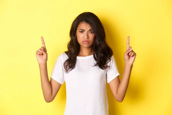 Portrait d'une adolescente afro-américaine bouleversée et déçue, se plaignant et fronçant les sourcils, pointant du doigt quelque chose de bouleversant, debout sur un fond jaune — Photo