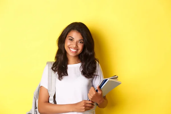 Portré boldog afro-amerikai női főiskolai hallgató, gazdaság notebook és hátizsák, mosolygós és áll a sárga háttér — Stock Fotó