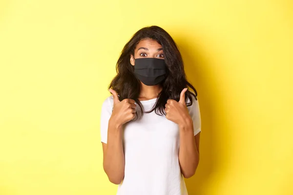 Il concetto di coronavirus, pandemia e stile di vita. Ritratto di bella ragazza afro-americana in maschera nera, mostrando pollici in alto in approvazione, come qualcosa di buono, sfondo giallo — Foto Stock
