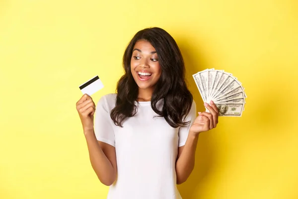 Portret van een aantrekkelijk Afrikaans-Amerikaans meisje, geld vasthoudend en kijkend naar een creditcard, staand over een gele achtergrond — Stockfoto