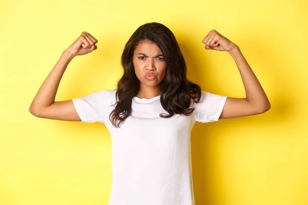 Imagen de una chica afroamericana fuerte y segura de sí misma, flexionando bíceps y pareciendo segura de sí misma, de pie sobre un fondo amarillo —  Fotos de Stock