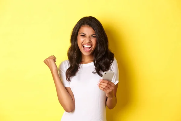 Bild av kaxig afrikansk-amerikansk flicka i vit t-shirt, gör knytnäven pump gest och hålla smartphone, firar prestation, står över gul bakgrund — Stockfoto