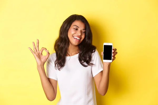 Retrato de linda chica afroamericana sonriendo complacido, mostrando el signo de bien y la pantalla del teléfono móvil, recomendando una aplicación o promoción, de pie sobre el fondo amarillo — Foto de Stock