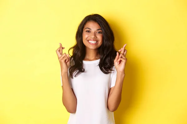 Retrato de chica afro-americana optimista y sonriente, cruzando los dedos para la buena suerte y pidiendo deseos, de pie sobre fondo amarillo — Foto de Stock