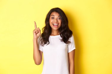 Heyecanlı gülümseyen Afrikalı Amerikalı kadının portresi. Bir fikri var, parmağını kaldırıyor ve önerisini söylüyor, sarı arka planda duruyor.