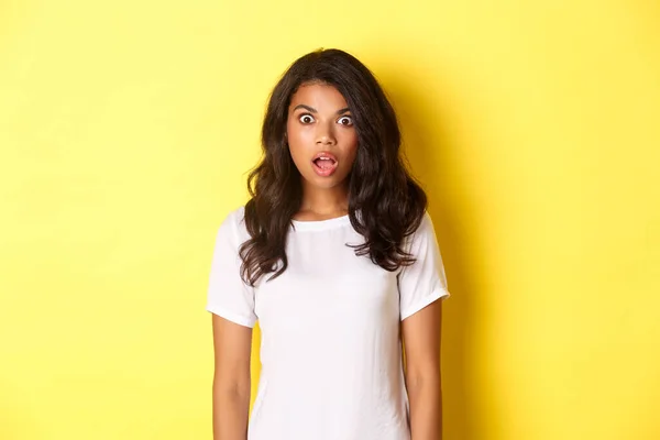Portret van geschokt en sprakeloos Afrikaans-Amerikaans meisje, kaak laten vallen en kijken naar geweldige promo aanbod, staande over gele achtergrond met kopieerruimte — Stockfoto