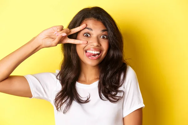 Imagem de bobo e bonito adolescente afro-americano menina, mostrando sinal de paz e sorrindo, de pé sobre fundo amarelo — Fotografia de Stock