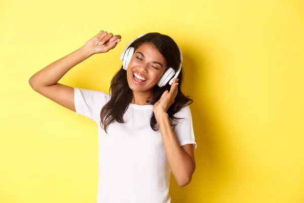 Jovencita alegre afroamericana escuchando música en auriculares, bailando alegre y cantando, de pie sobre fondo amarillo — Foto de Stock