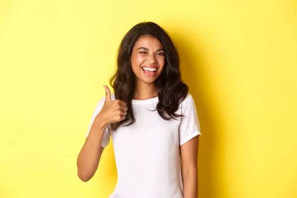 Retrato de una estudiante afro-americana feliz y satisfecha, mostrando pulgares hacia arriba y guiños, sonriendo complacida, aprobar algo bueno, alabándote, de pie sobre el fondo amarillo — Foto de Stock