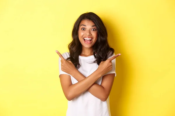 Porträt einer glücklichen und erstaunten afrikanisch-amerikanischen Frau, die zwei Promoangebote zeigt, die Finger zur Seite zeigt und aufgeregt lächelt, vor gelbem Hintergrund stehend — Stockfoto