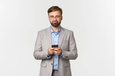 İş elbisesi ve gözlüklü, hayal kırıklığına uğramış sakallı bir adamın portresi, elinde akıllı telefon, üzgün görünüyor, beyaz arka planda duruyor.