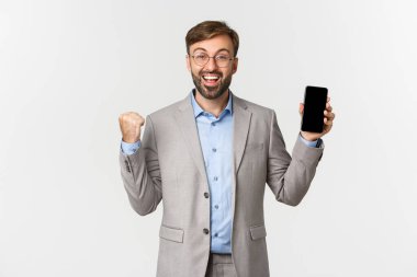 Gri takım elbiseli ve gözlüklü mutlu işadamı, mobil uygulamalarda günlük hedefe ulaşır, akıllı telefon ekranını gösterir ve sevinir, bir şeyler kazanır, beyaz arka planda durur.