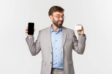 Sakallı, gri takım elbiseli ve gözlüklü neşeli erkek girişimci cep telefonu ekranında ve kredi kartında uygulama gösteriyor, heyecanlı bir şekilde gülümsüyor, beyaz arka planda duruyor.