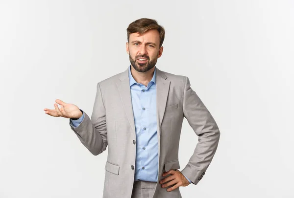 Ritratto di bell'uomo d'affari barbuto in abito grigio, alzando la mano e accigliandosi, guardando confuso, in piedi su sfondo bianco — Foto Stock