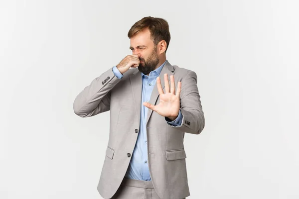 Retrato de empresário barbudo nojento em terno cinza, rejeitando algo com cheiro horrível, fechar o nariz e fazer sinal de declínio, em pé sobre fundo branco — Fotografia de Stock