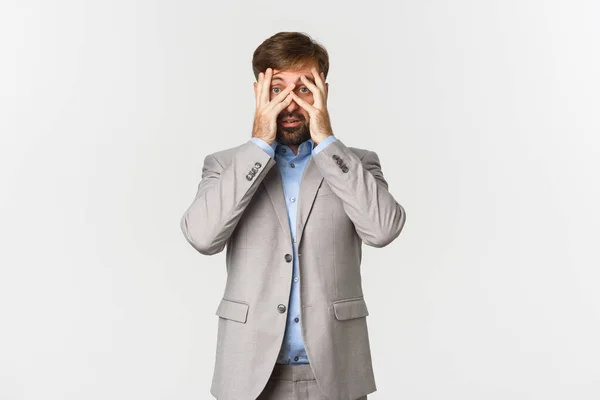 Väijytetyn liikemiehen muotokuva harmaassa puvussa, peittävät silmät ja kurkistavat sormien läpi pelokkailla kasvoilla, valkoisella taustalla — kuvapankkivalokuva