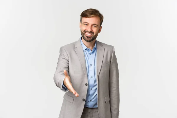 Porträtt av leende och självsäker skäggig affärsman, sträcker handen för handslag, hälsning affärspartner, står i grå kostym över vit bakgrund — Stockfoto