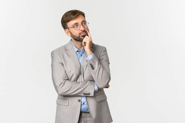 Retrato de homem de negócios pensativo com barba, vestindo terno cinza e óculos, pensando em algo com rosto sério, tomando decisão, de pé sobre fundo branco — Fotografia de Stock