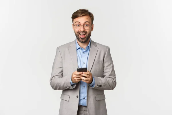 Retrato de empresário bonito com barba, vestindo terno cinza e óculos, olhando feliz e sorridente depois de ler algo interessante no telefone celular, em pé sobre fundo branco — Fotografia de Stock