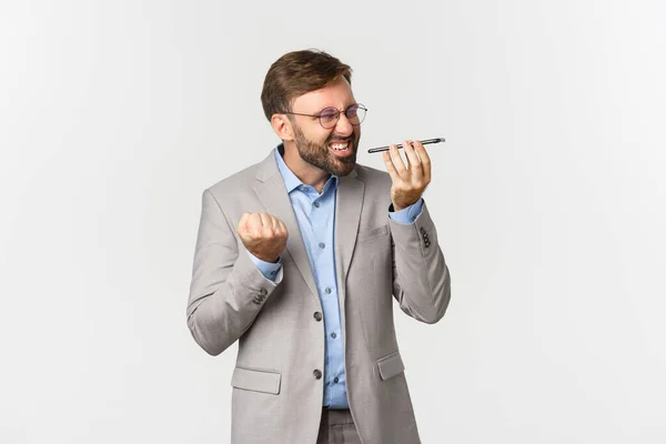 Pissed-off and angry businessman with beard, wearing grey suit and glasses, shouting at speakerphone with mad expression, standing over white background — Fotografia de Stock