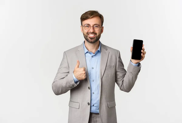 Hezký usměvavý podnikatel s vousy, v šedém obleku a brýlích, ukazující palec nahoru a předvádějící něco na mobilním telefonu, doporučujeme aplikaci, bílé pozadí — Stock fotografie