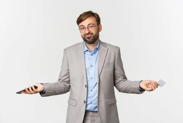 Image of clueless smiling businessman in glasses and gray suit, spread hands sideways and shrugging confused, holding mobile phone and credit card — Stock Photo, Image