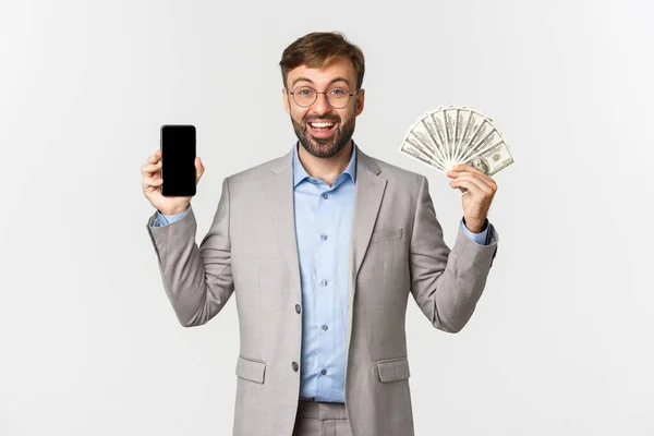 Hombre de negocios exitoso en gafas y traje gris, mostrando la pantalla del teléfono inteligente y el dinero, sonriendo emocionado, de pie sobre el fondo blanco — Foto de Stock