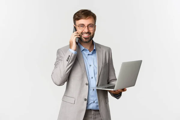 Beeld van een succesvolle en zelfverzekerde zakenman in een bril en een grijs pak, die telefoneert en een laptop vasthoudt, blij lacht op de camera, over een witte achtergrond staat — Stockfoto