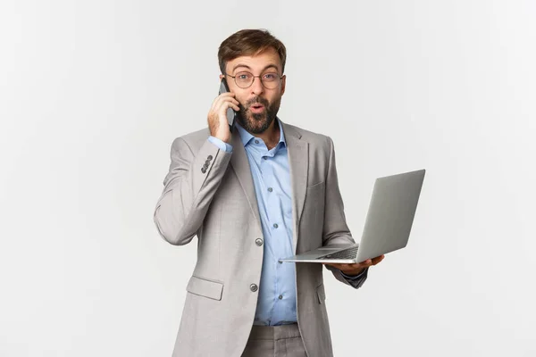 Portret van knappe zakenman in grijs pak en bril, goed nieuws ontvangen tijdens telefoongesprek, laptop vasthouden, tevreden kijken, over een witte achtergrond staan — Stockfoto