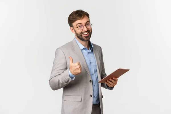 Портрет счастливого бизнесмена в сером костюме и очках, хваля хорошую работу, читая что-то на цифровой планшете и показывая большой палец вверх в одобрении, стоя на белом фоне — стоковое фото