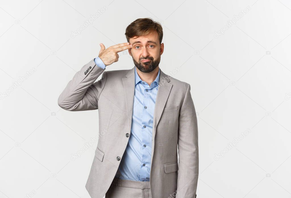 Image of miserable businessman with beard, wearing grey suit, making finger gun sign over head and shooting himself, standing distressed over white background
