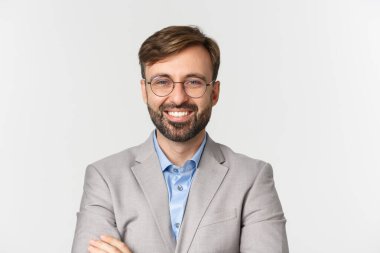 Kendine güvenen, yakışıklı, patronluk taslayan, gözlüklü, gri takım elbiseli, göğsünde çapraz kolları olan ve gülümseyen, beyaz arka planda duran bir erkek.