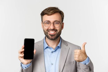 Gözlüklü ve gri takım elbiseli yakışıklı erkek girişimcinin yakın çekimi, cep telefonu ekranı ve onaylı baş parmak gösterimi, uygulama önerisi, beyaz arkaplan üzerinde durmak