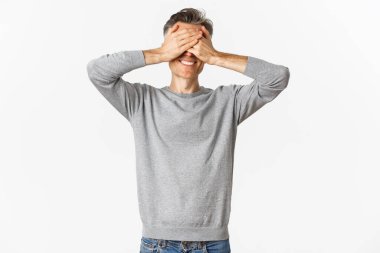 Yakışıklı, mutlu, orta yaşlı bir adamın portresi şaşırmayı bekliyor, gözleri kapalı, gülümsüyor, saklambaç oynuyor, beyaz arka planda duruyor.