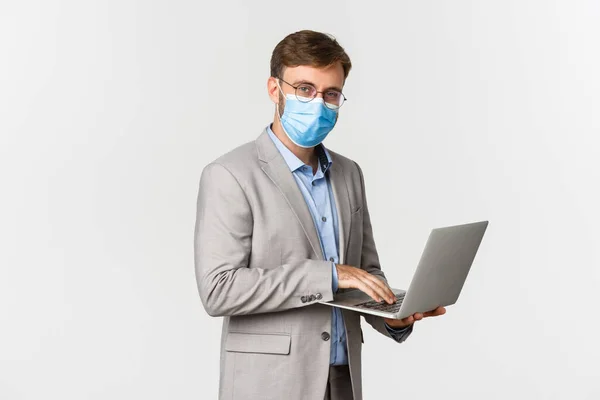 Begreppet arbete, covid-19 och social distansering. Bild av arbetande affärsman i medicinsk mask och kostym, med bärbar dator och leende glad, står över vit bakgrund — Stockfoto