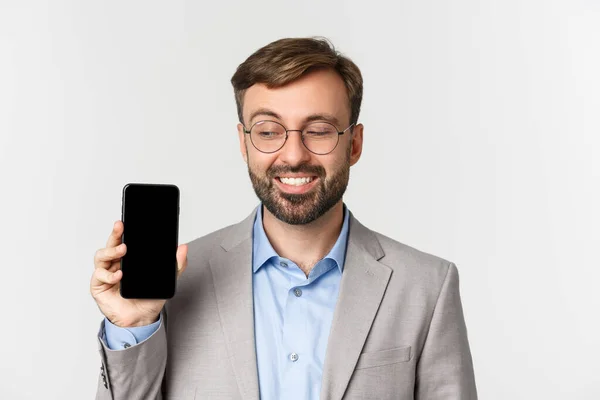 Närbild av stilig skäggig affärsman i grå kostym och glasögon, visar mobiltelefon och ser stolt, leende nöjd och står över vit bakgrund — Stockfoto