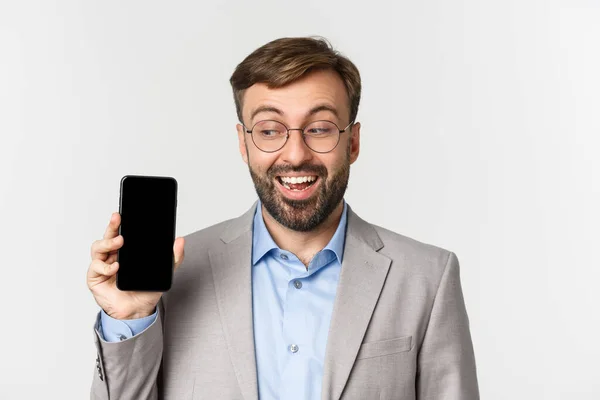 Närbild av upphetsad affärsman i grå kostym och glasögon, visar mobiltelefon skärmen och ser glad, står över vit bakgrund — Stockfoto