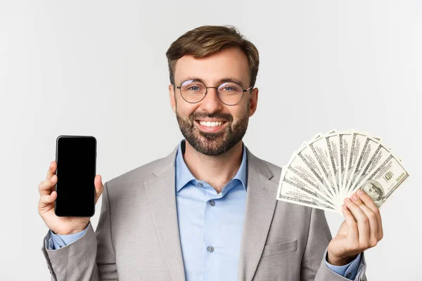 Primer plano del exitoso hombre de negocios en traje gris y gafas, mostrando el dinero y la pantalla del teléfono inteligente, de pie sobre fondo blanco — Foto de Stock