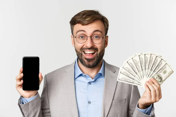 Primer plano del exitoso hombre de negocios en traje gris y gafas, mostrando el dinero y la pantalla del teléfono inteligente, sonriendo alegre, de pie sobre fondo blanco — Foto de Stock