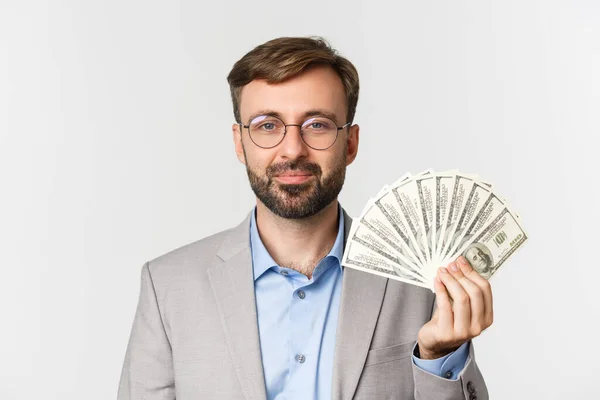Zbliżenie męskiego przedsiębiorcy w szarym garniturze i okularach uśmiechniętych, pokazujących pieniądze, stojących na białym tle — Zdjęcie stockowe