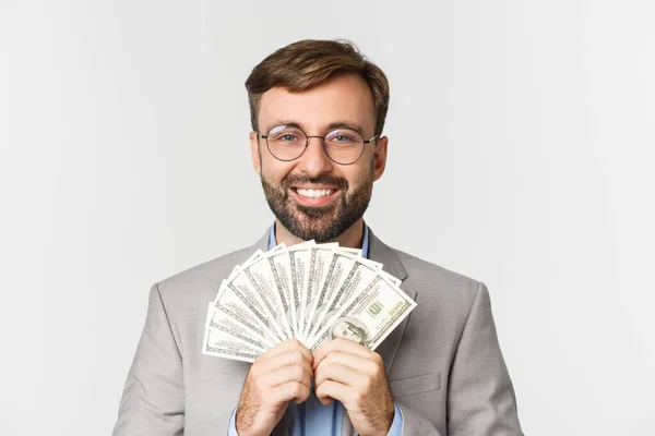 Primer plano del hombre de negocios rico y exitoso, con traje gris y gafas, mostrando dinero en efectivo y sonriendo feliz, de pie sobre fondo blanco — Foto de Stock