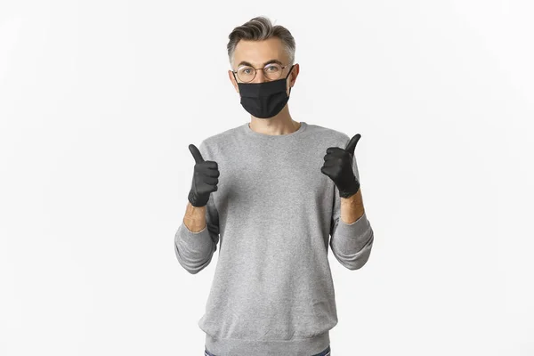 Covid-19, pandemic and social distance concept. Изображение мужчины средних лет в черной медицинской маске, перчатках и очках, показывающих большие пальцы вверх, защищая себя от заражения коронавирусом — стоковое фото