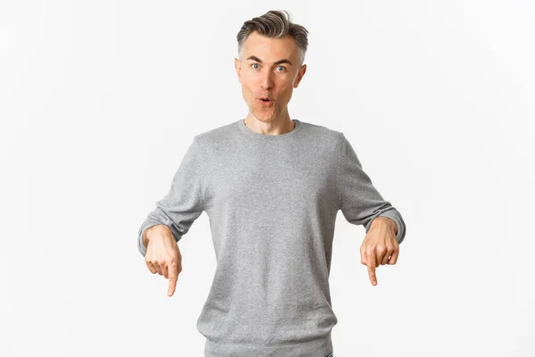 Здивований красивий хлопець середнього віку в сірому светрі ставить питання про продукт, вказуючи пальці вниз і дивуючись, стоїть над білим тлом — стокове фото