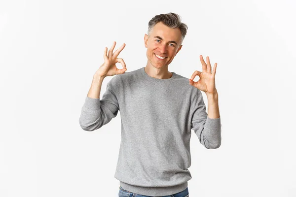 Retrato de homem bonito bem sucedido em camisola cinza, mostrando sinais ok e sorrindo satisfeito, aprovar algo bom, recomendando produto, de pé sobre fundo branco — Fotografia de Stock