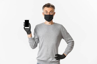 Covid-19 kavramı, sosyal mesafeler ve yaşam tarzı. Şaşırmış orta yaşlı bir adamın görüntüsü internette bir şey buldu. Akıllı telefon ekranını gösteriyor ve şaşırmış görünüyor. Eldivenli tıbbi maske takıyor.