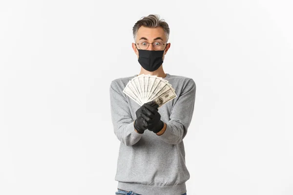 Concepto de covidio-19, distanciamiento social y estilo de vida. Guapo hombre de mediana edad sorprendido en gafas, máscara médica y guantes, mostrando el dinero del premio, ganar la lotería, de pie sobre fondo blanco —  Fotos de Stock