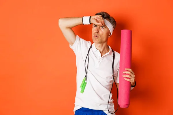 Concept of workout, gym and lifestyle. Image of handsome middle-aged man, tired after fitness exercises, holding skipping rope and yoga mat, wiping sweat off forehead, orange background