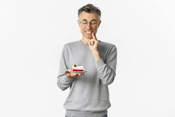 Porträt eines gutaussehenden Mannes mittleren Alters mit Brille und grauem Pullover, der verführt auf leckeren Kuchen schaut, Dessert essen will, vor weißem Hintergrund steht — Stockfoto